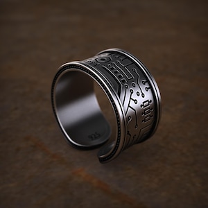 Peut inclure: Une bague argentée avec un motif de circuit imprimé. La bague a un design ouvert et présente des lignes gravées et des détails circulaires, créant une esthétique inspirée de la technologie. La bague est présentée sur une surface marron.
