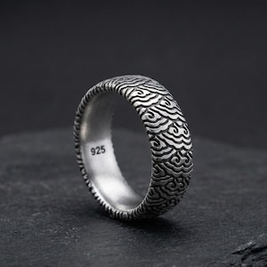 May include: A silver ring with a wave-like pattern. The ring is made of sterling silver and has the number "925" stamped on the inside. The ring is on a dark gray surface.