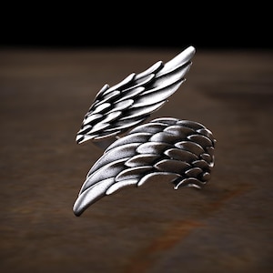 May include: A silver ring with a wing design. The ring features two detailed wings, one on each side, with textured feather detailing. The wings are slightly curved, giving a sense of movement. The ring is set against a dark background.