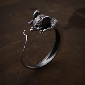 May include: A silver ring featuring a detailed mouse design. The mouse is depicted with textured fur and large ears, curving around the finger. The ring is made of polished metal, with the hallmark "925" visible on the band.