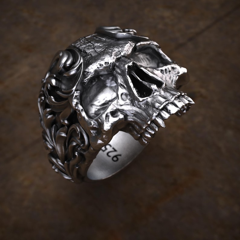 May include: A detailed silver skull ring with intricate carvings and a textured finish. The ring features a skull design with visible teeth and eye sockets, and decorative floral patterns on the sides. The band is marked with "925".