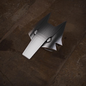 May include: A silver-colored ring with a geometric design. The ring features a central, elongated shape with a pointed end, flanked by two triangular projections. The ring's surface has a brushed metal texture, and it is set against a brown background.