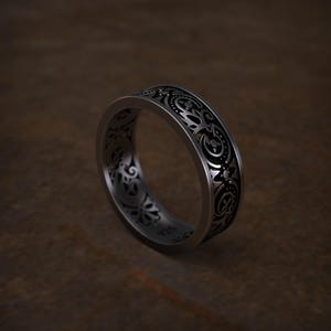 May include: A silver ring with an intricate, repeating floral and scrollwork pattern. The ring has a dark background within the design, creating contrast. The band is approximately 0.25 inches wide. The ring is set against a brown background.