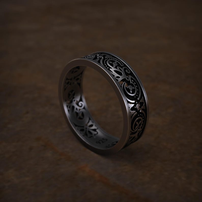 May include: A silver ring with an intricate, repeating floral and scrollwork pattern. The ring has a dark background within the design, creating contrast. The band is approximately 0.25 inches wide. The ring is set against a brown background.