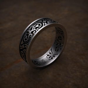 May include: A silver ring with an intricate black floral pattern. The ring has a wide band with detailed engravings. The design includes swirling lines, dots, and floral motifs. The ring is set against a dark brown background.