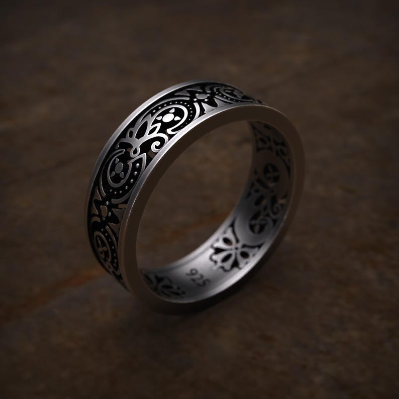 May include: A silver ring with an intricate black floral pattern. The ring has a wide band with detailed engravings. The design includes swirling lines, dots, and floral motifs. The ring is set against a dark brown background.