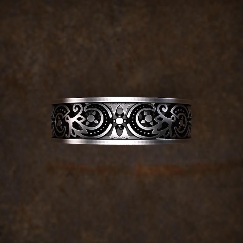 May include: A silver ring with an intricate floral and geometric pattern. The band features a repeating design of stylized flowers and circular elements, creating a detailed and ornamental appearance. The ring is displayed against a dark brown background.