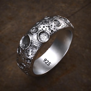 Könnte beinhalten: Ein silberner Ring mit strukturierter Oberfläche, die der Mondoberfläche mit Kratern ähnelt. Der Ring ist innen mit "925" markiert. Der Ring ist vor einem braunen Hintergrund platziert.