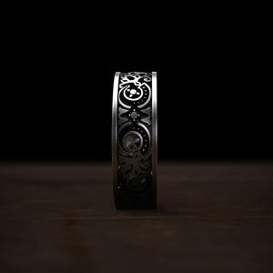 May include: A silver ring with an intricate, repeating pattern of floral and geometric designs. The ring's surface is detailed with dark accents, creating a contrast against the polished metal. The ring is displayed on a dark wooden surface.