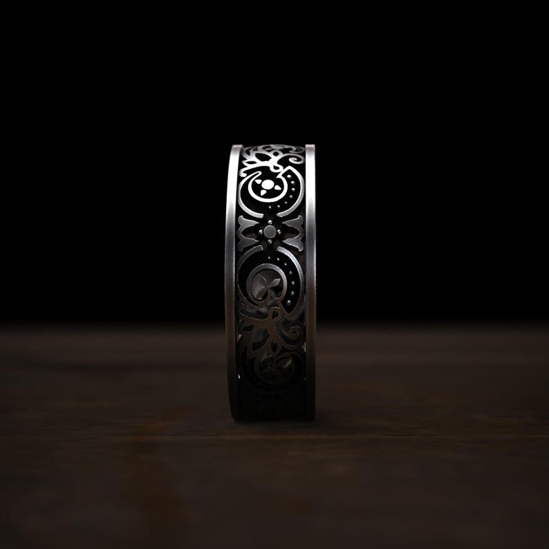 May include: A silver ring with an intricate, repeating pattern of floral and geometric designs. The ring's surface is detailed with dark accents, creating a contrast against the polished metal. The ring is displayed on a dark wooden surface.