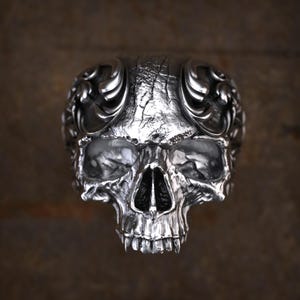 May include: A detailed, silver-colored skull ring with intricate carvings. The ring features a realistic skull design with detailed eye sockets, nose, and teeth. The top of the ring has decorative flourishes. The metal has a textured, aged appearance.