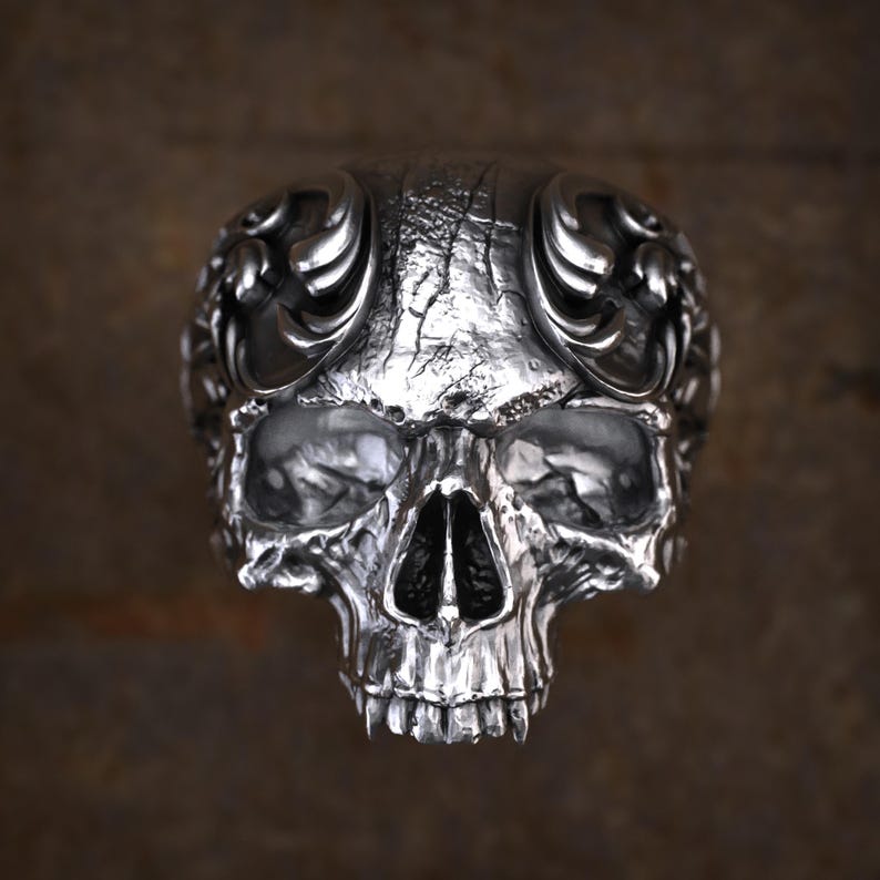 May include: A detailed, silver-colored skull ring with intricate carvings. The ring features a realistic skull design with detailed eye sockets, nose, and teeth. The top of the ring has decorative flourishes. The metal has a textured, aged appearance.