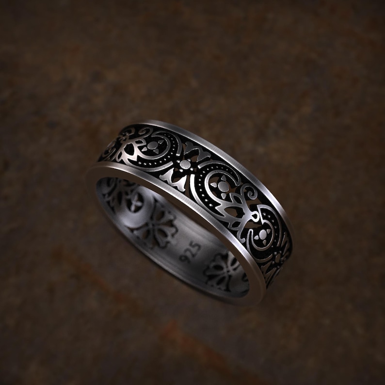 May include: A silver ring with an intricate, openwork design. The band features a repeating pattern of swirling shapes and floral elements. The ring is marked with "925" on the inside, indicating its silver content. The ring is displayed against a brown background.