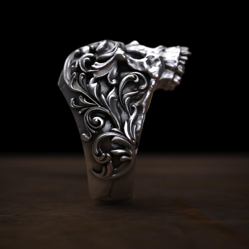 May include: A silver ring with a skull design. The ring features intricate floral scrollwork on the band and the skull's forehead. The skull's teeth are visible. The ring is set against a dark background.