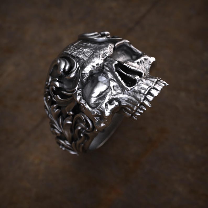 May include: A silver skull ring with intricate details. The ring features a detailed skull design with visible teeth and eye sockets. The sides of the ring are adorned with ornate, swirling patterns. The ring is made of a shiny, metallic material.