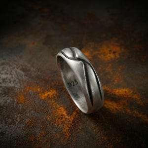 May include: A silver ring with a textured design and the number "925" engraved on the inside. The ring has a matte finish and a unique, flowing pattern. The ring is displayed against a dark, textured background.