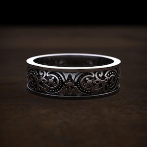 May include: A silver-colored ring with an intricate, repeating floral and scrollwork pattern. The band is approximately 0.25 inches wide. The design features detailed, symmetrical elements, creating a vintage or antique aesthetic. The ring is displayed on a dark wooden surface.