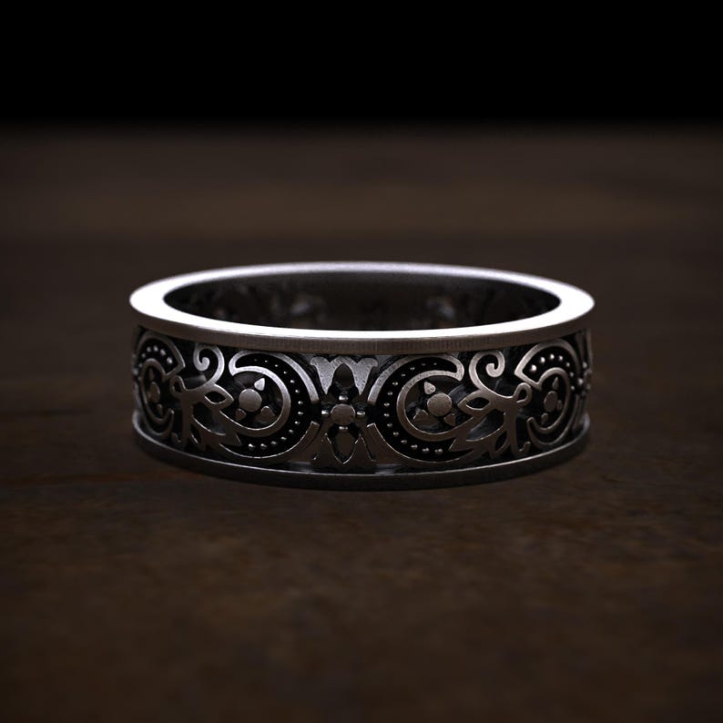May include: A silver-colored ring with an intricate, repeating floral and scrollwork pattern. The band is approximately 0.25 inches wide. The design features detailed, symmetrical elements, creating a vintage or antique aesthetic. The ring is displayed on a dark wooden surface.