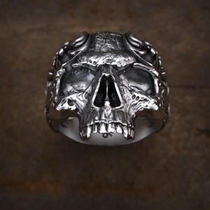May include: A silver skull ring with detailed facial features, including eye sockets, a nose cavity, and teeth. The ring has a textured surface and a dark background, creating a gothic or biker aesthetic.