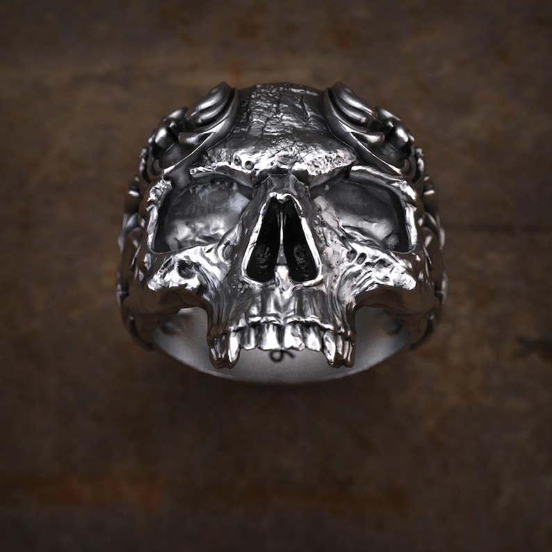 May include: A silver skull ring with detailed facial features, including eye sockets, a nose cavity, and teeth. The ring has a textured surface and a dark background, creating a gothic or biker aesthetic.