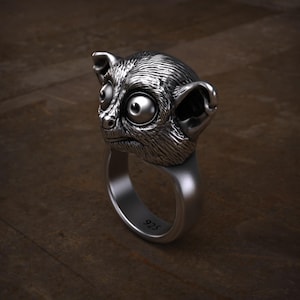 Könnte beinhalten: Ein silberner Ring mit einem detaillierten Primatenkopf, der große Augen und markante Ohren aufweist. Der Ring besteht aus poliertem Metall und ist vor einem dunklen Holzgrund platziert. Der Ring ist mit "925" markiert.