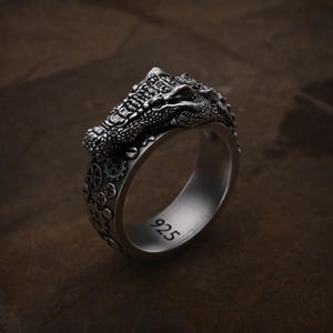 May include: A silver ring featuring a detailed crocodile head design. The ring has a textured surface and is engraved with the number "925". The band is decorated with gear-like patterns, creating a unique and intricate accessory.