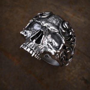 May include: A detailed silver skull ring with intricate carvings. The ring features a realistic skull design with visible teeth, eye sockets, and textured surfaces. The ring is a bold statement piece, perfect for those who appreciate gothic or biker-inspired jewelry.