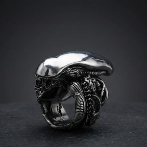 Könnte beinhalten: Ein silberner Ring mit detaillierter Darstellung des Kopfes eines Alien-Kreatur. Der Ring zeigt den länglichen Kopf, das strukturierte Exoskelett und die bedrohlichen Gesichtszüge der Kreatur. Der Ring ist vor einem dunklen Hintergrund platziert.
