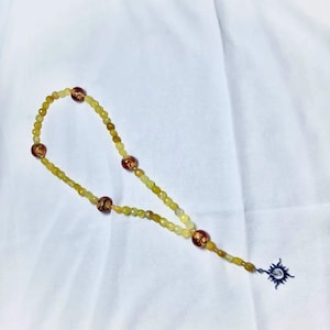 Può includere: Una collana di perline gialle e marroni con un ciondolo d'argento all'estremità. Il ciondolo è un sole stilizzato con raggi.