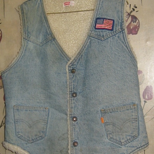 May include: A light blue denim vest with a white sherpa lining, featuring a button-front closure, two front pockets with stitched detailing, and a small American flag patch on the left chest. A Levi's brand label is visible at the inner neckline.