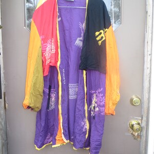 May include: A colorful, patterned robe with a mix of vibrant colors, including purple, orange, red, yellow, and black. The robe features unique designs and is hanging on a wire hanger. The robe is made of a lightweight fabric.