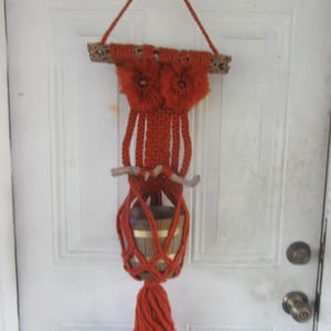 May include: A handmade macrame owl plant hanger in shades of orange. The owl features a wooden dowel, two fluffy flower-like eyes, and a woven body that holds a small wooden planter. A tassel hangs from the bottom.