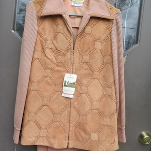 May include: A tan suede jacket with a geometric design, featuring a zip-up front and light pink collar and sleeves. The jacket has a tag that reads "PALENA KNITWEAR".