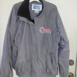 Può includere: Giacca grigia con cerniera, fodera in pile nero e logo Main Gate sul petto sinistro. La giacca ha un logo "Major Jones Racing" sul davanti.