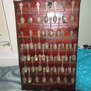 May include: A wooden spoon rack with multiple silver-colored spoons. The rack is dark brown with a decorative top. Each spoon has a detailed handle and a small bowl. The spoons are arranged in rows, creating a display.