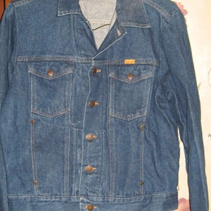 May include: A medium-wash denim jacket with a classic design. Features a collared neckline, button-up front, and two chest pockets. The jacket has a straight hem and is made of durable denim material. The brand name "RUSTLER" is visible on the chest pocket.