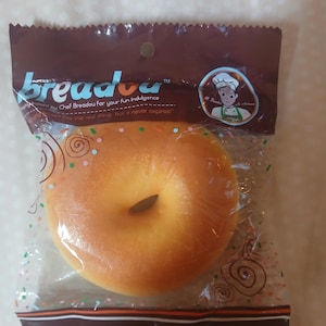 May include: A packaged, orange-yellow bagel-shaped squishy toy. The clear plastic packaging has the text "breadou" and "Chef Breadou for your Fun Indulgence" printed on it. The packaging is sealed with a brown top and bottom border.