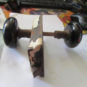 May include: An antique door knob set with black, oval-shaped knobs and a rusty, rectangular lock mechanism. The metal components show signs of age with peeling white paint and a weathered appearance. The set includes a spindle and is likely for a vintage door.