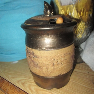 May include: A ceramic honey pot with a brown and tan glaze. The pot has a lid with a small animal figurine on top. The pot has a wooden dipper.