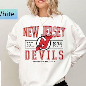 Könnte beinhalten: Weißes Sweatshirt mit dem Text "NEW JERSEY DEVILS" in Rot und einem schwarz-roten Logo. Der Text "EST. 1974" ist ebenfalls vorhanden. Die Worte "NATIONAL HOCKEY LEAGUE" stehen unten. Das Sweatshirt hat einen Rundhalsausschnitt.