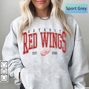 May include: Sport Grey sweatshirt featuring the Detroit Red Wings logo in red. The design includes the team name, a winged wheel logo, and the year 1926. The sweatshirt is a light gray color.