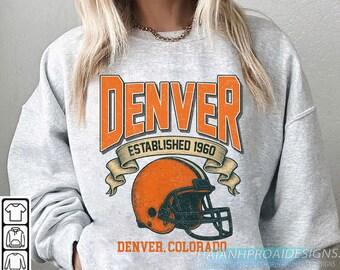 May include: A light grey sweatshirt featuring a distressed orange graphic. The graphic reads "DENVER ESTABLISHED 1960" above an orange American football helmet. Below the helmet is "DENVER, COLORADO". The sweatshirt has a crew neck.