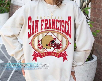 May include: Cream-coloured sweatshirt with a distressed red and gold graphic. The graphic features the words "FOOTBALL SAN FRANCISCO" above an oval with a football helmet and the text "EST. 1946". The sweatshirt has long sleeves.