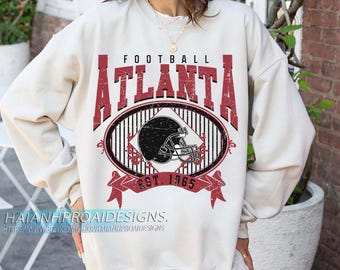 Puede incluir: Sudadera color crema con un gráfico rojo y negro desgastado. El diseño presenta la palabra "ATLANTA" en letras en negrita, la palabra "FOOTBALL", un casco de fútbol y el texto "EST. 1965".