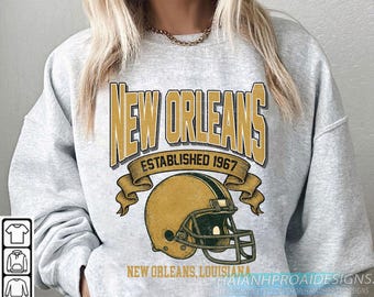 May include: A light gray sweatshirt with a gold and black graphic. The graphic reads "NEW ORLEANS ESTABLISHED 1967" above a football helmet. Below the helmet, it says "NEW ORLEANS, LOUISIANA".