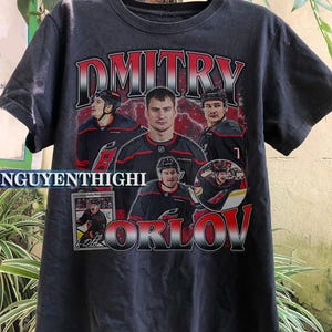 May include: Dark gray t-shirt featuring a hockey player graphic. The shirt displays the name "Dmitry Orlov" in red and white lettering, along with multiple images of the player in action. The shirt also includes the text "NGUYENTHIGHI".