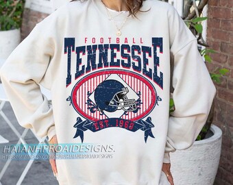 Op de afbeelding: Crèmekleurige sweatshirt met een distressed grafisch ontwerp. Het ontwerp toont het woord "TENNESSEE" in marineblauwe blokletters boven een ovaal met een footballhelm en de tekst "EST. 1969". De sweatshirt heeft lange mouwen.