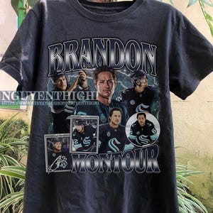 May include: Black t-shirt with a graphic of hockey player Brandon Montour. The graphic features a collage of photos of Montour in his hockey uniform, along with his name and signature.