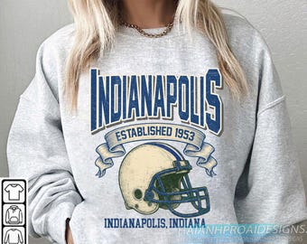 May include: A light gray sweatshirt featuring a blue and white graphic of "Indianapolis" above a football helmet. The text "Established 1953" and "Indianapolis, Indiana" are also displayed. The design has a vintage, collegiate aesthetic.