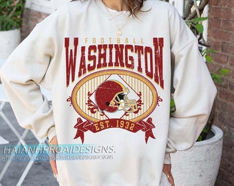 Op de afbeelding: Crèmekleurige sweatshirt met een verweerde bordeauxrode en gouden afbeelding. De afbeelding toont het woord "WASHINGTON" boven een ovaal met een voetbalhelm en de tekst "EST. 1932". De sweatshirt heeft lange mouwen.